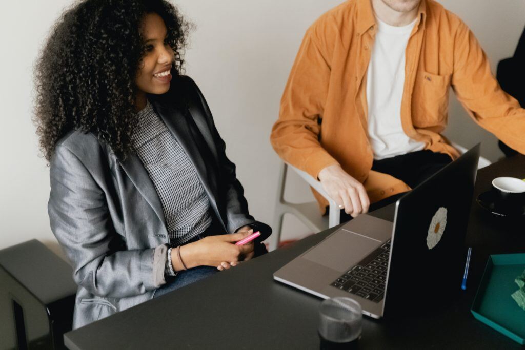 A Black woman with long curly hair wearing a dark gray blazer and checked blouse sits in front of a laptop atop a black conference table. She is smiling. A white man wearing an open orange shirt over a white T-shirt sits to her left, and his head is cut out of the frame.