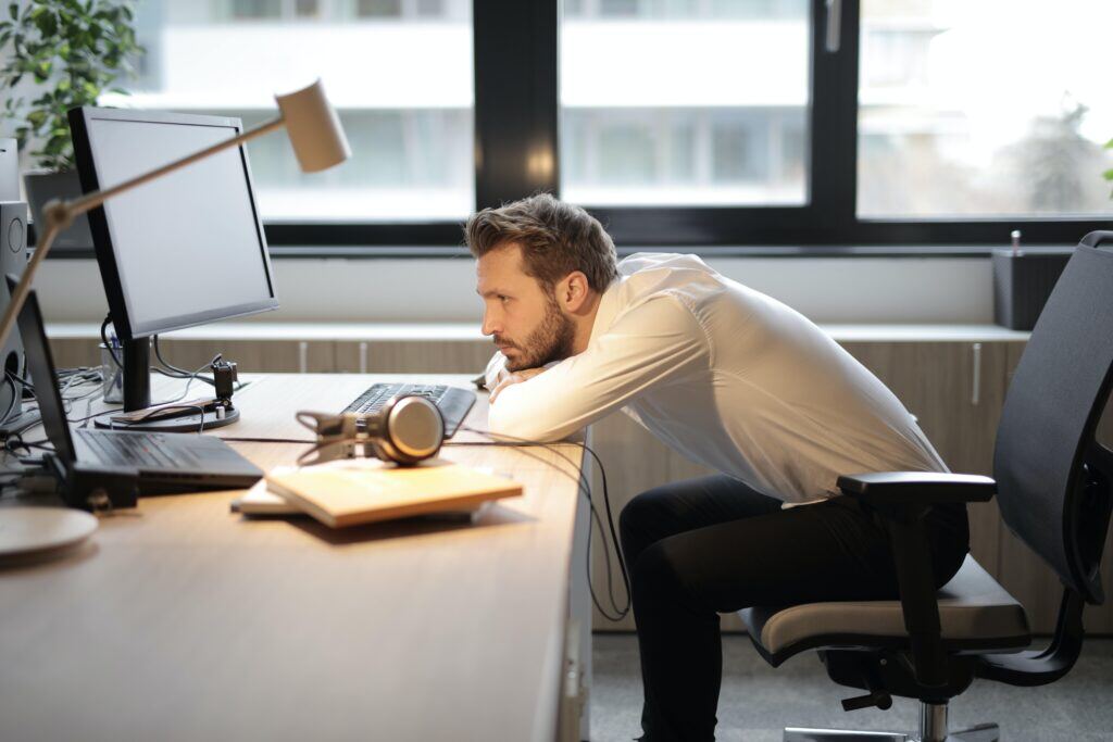 A pale-skinned man with short hair and a beard, wearing a white button-down shirt and black pants, is sitting in a dimly lit office environment. The desk holds a laptop, a monitor, a task lamp, a teetering stack of books, an empty dinner plate, and a pair of headphones. He is sitting in a black office chair and leaning over his desk, resting his chin on his arms. His expression is dull, and he appears to be checked out—his disengagement could be quiet quitting or perhaps burnout.