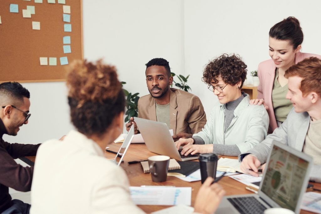 Hiring Gen Z is essential to surviving in this talent market. In this photo, a multiracial, multiethnic, and gender-diverse group of Gen Z professionals sit around a conference table in business casual attire. Some of the Gen Z workers are using laptops. Other Gen Z employees are looking at others’ laptop displays. The room has white walls, a corkboard with sticky notes, a leafy plant in the corner, and diffuse bright light. The conference table is cluttered with coffee mugs, notebooks, writing utensils, and various documents.