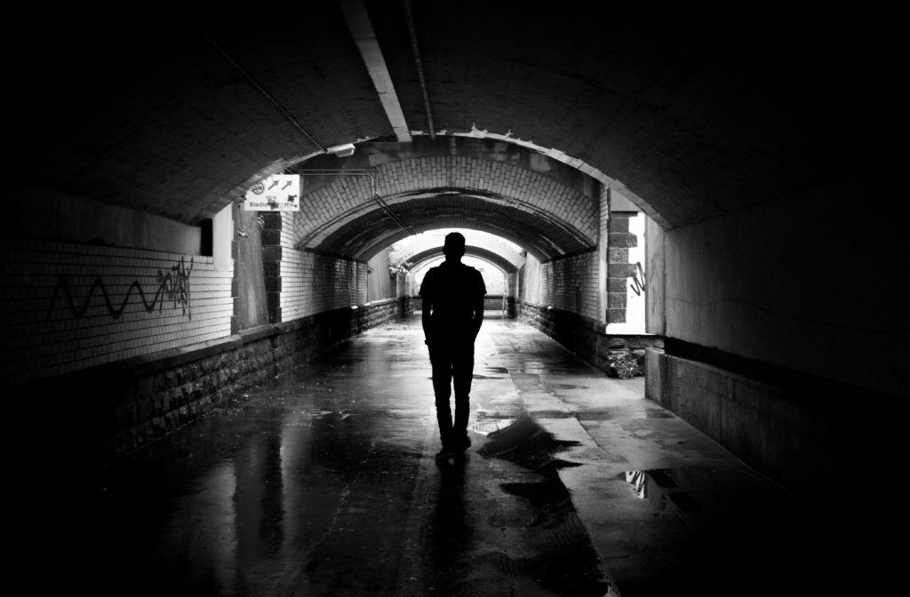 A silhouette of a man standing in a dark, damp, graffitied tunnel. The photo is in black and white, and its dark, gritty mood evokes the theme of the psychology of criminal behavior. The man’s anonymity represents the fact that, historically, little has been known about the role of personality in organized crime.
